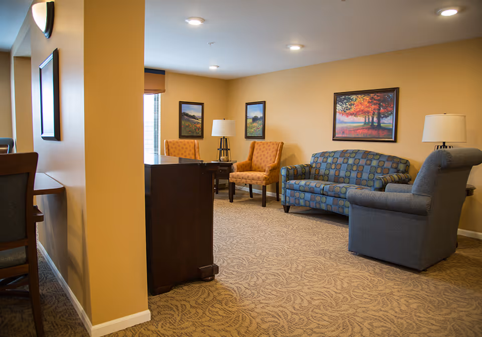 A cozy senior living common area with beige walls and patterned carpet. The room features a blue patterned sofa, a gray armchair, two orange armchairs, two table lamps, and framed landscape paintings on the walls. A dark wooden desk is partially visible near the center.