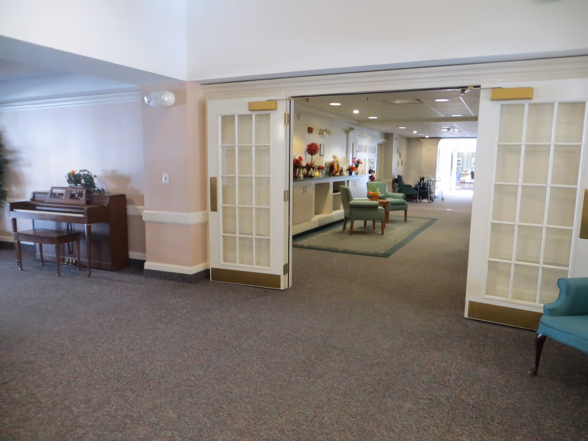 Interior view of a senior living community common area with open double glass doors leading to a hallway. The hallway has chairs, a carpet, and decorative items on a sideboard. A piano with a bench and a plant is visible on the left side of the image.