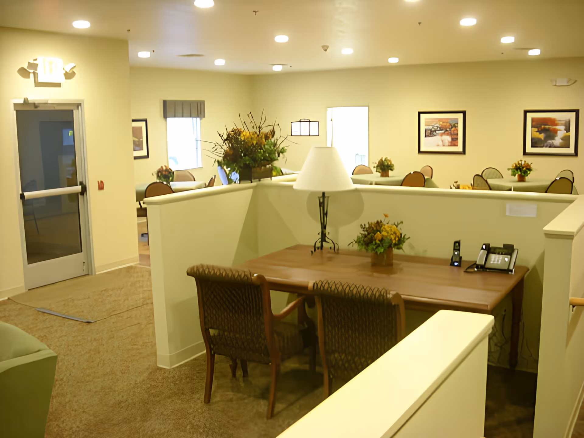 Interior view of a senior living facility common area with a wooden table, two chairs, a lamp, and a flower arrangement in the foreground. In the background, there are multiple tables and chairs with flower centerpieces, framed artwork on the walls, and a door leading to another room.
