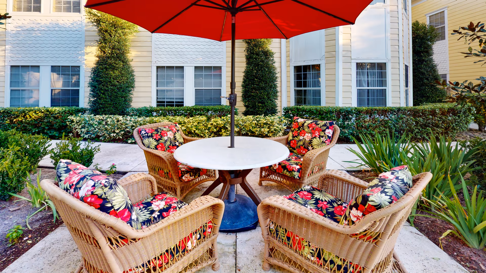 Wicker patio chairs with floral cushions around a round table under a red umbrella outside a yellow building.