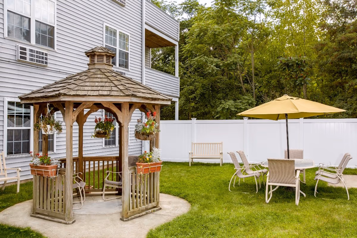 Outdoor area at The Village at Kensington Place featuring a wooden gazebo with hanging flower baskets and potted plants, a round table with six chairs under a yellow umbrella, a wooden bench, green grass, white privacy fence, and trees in the background.
