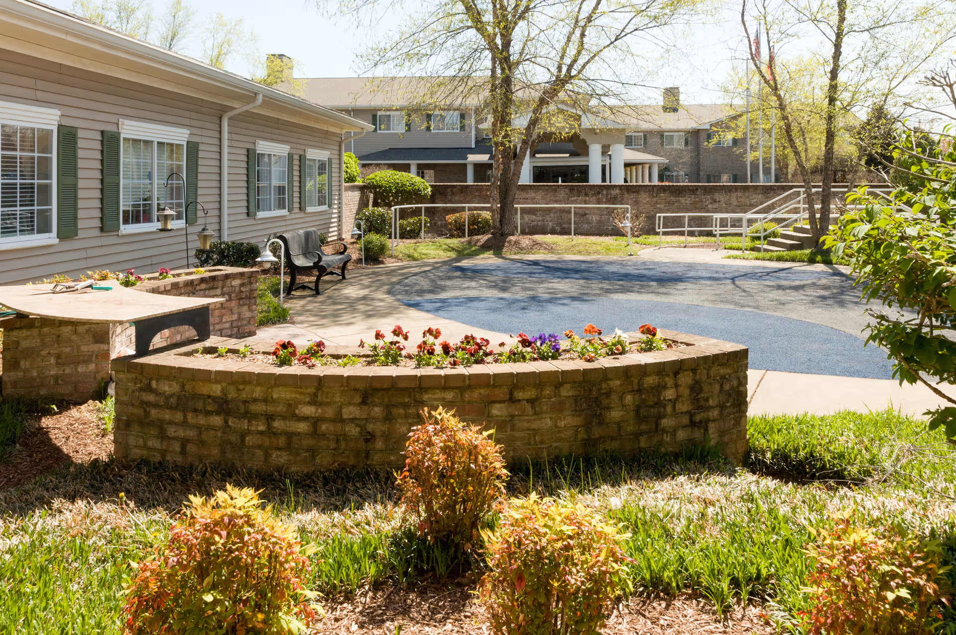 Outdoor garden area at a senior living facility with a curved brick flower bed filled with colorful flowers, a bench along the building wall, and a paved area with a ramp and stairs. Trees and shrubs surround the space under a clear sky.