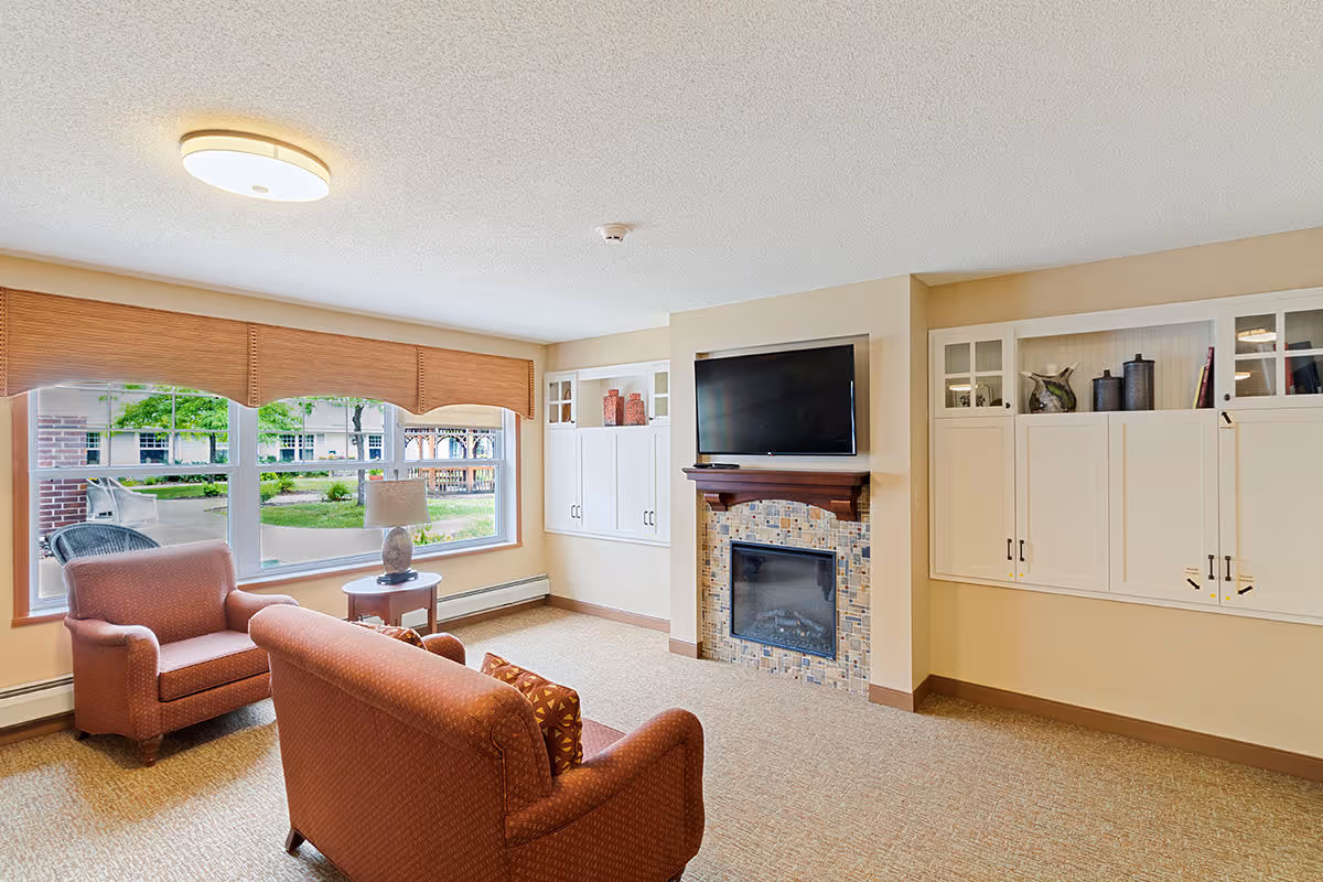A cozy senior living room with a large window featuring a view of the outdoor garden area. The room has a comfortable armchair and loveseat in a reddish pattern, a small round side table with a lamp, a fireplace with a tiled surround, and a flat-screen TV mounted above it. Built-in white cabinets with glass doors flank the fireplace.