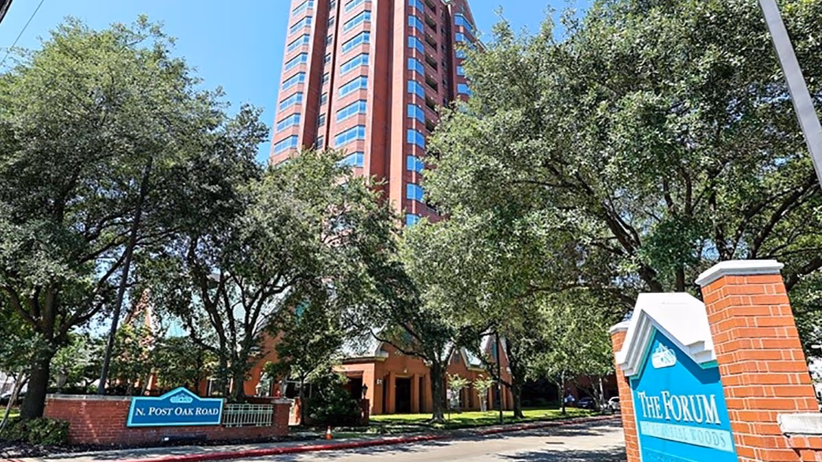 Exterior view of a tall red-brick building partially obscured by large green trees under a clear blue sky. In the foreground, there is a sign that reads 'The Forum at Memorial Woods' and another sign indicating 'N. Post Oak Road'.