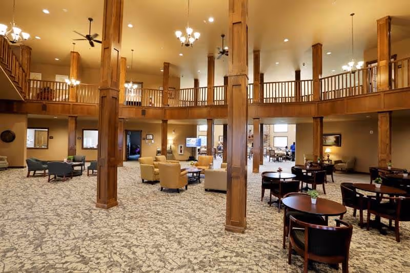Spacious senior living facility common area with high ceilings, wooden pillars, and a mezzanine level. The room is furnished with multiple seating arrangements including armchairs, sofas, and round tables with chairs. The carpet has a patterned design, and the lighting includes chandeliers and ceiling fans.