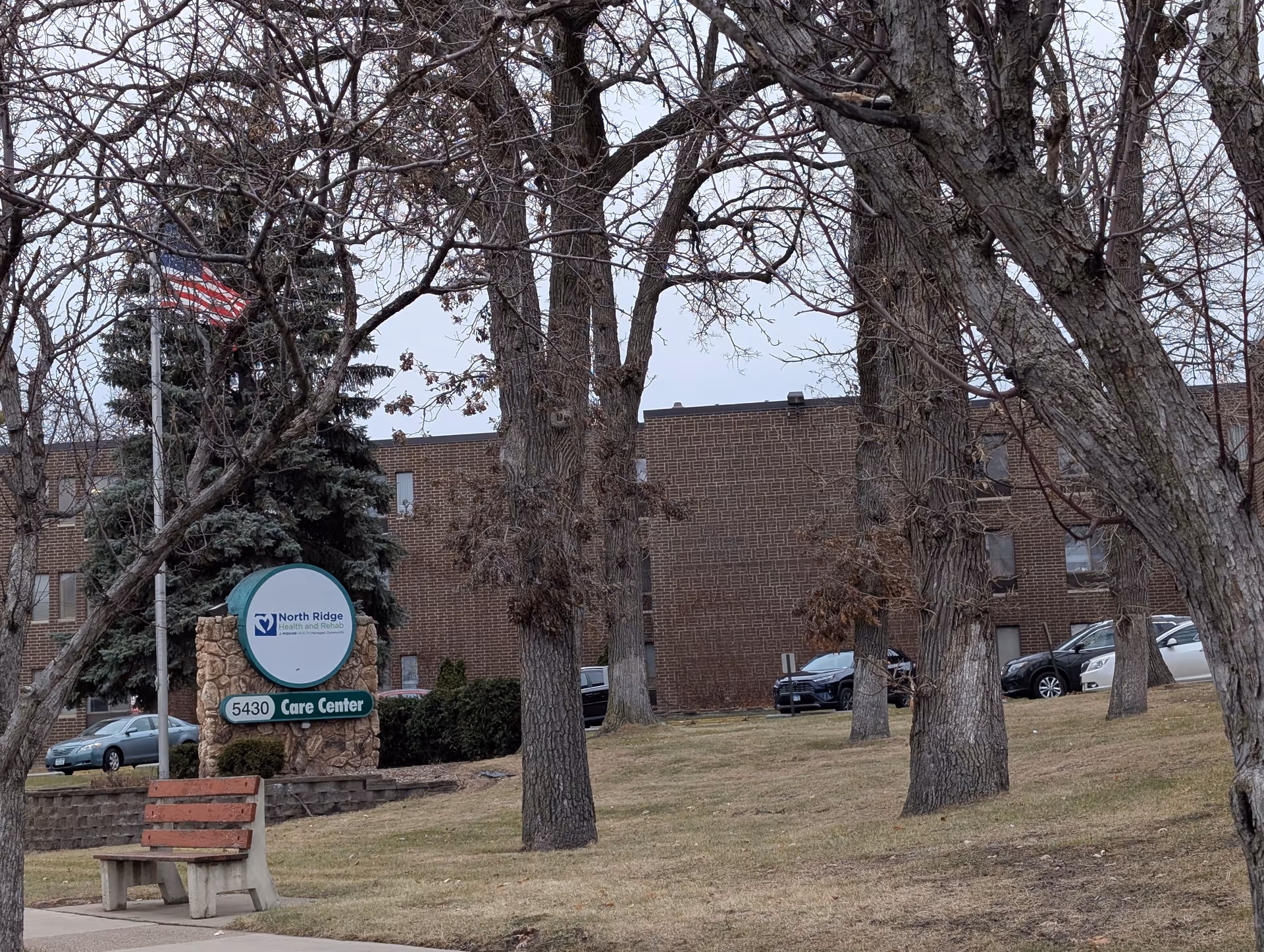 Front exterior of a brick senior care center with a North Ridge sign, leafless trees, a bench, and parked cars.