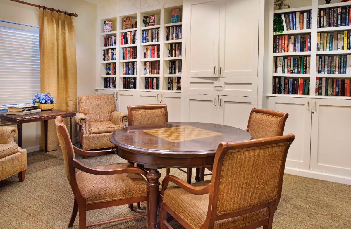 A cozy room with a round wooden table featuring a built-in chessboard, surrounded by four cushioned wooden chairs. Behind the table are built-in white bookshelves filled with books and closed cabinets. To the left, there are two upholstered armchairs next to a small wooden table with a basket of blue flowers and some books. A window with closed blinds and beige curtains is also visible.
