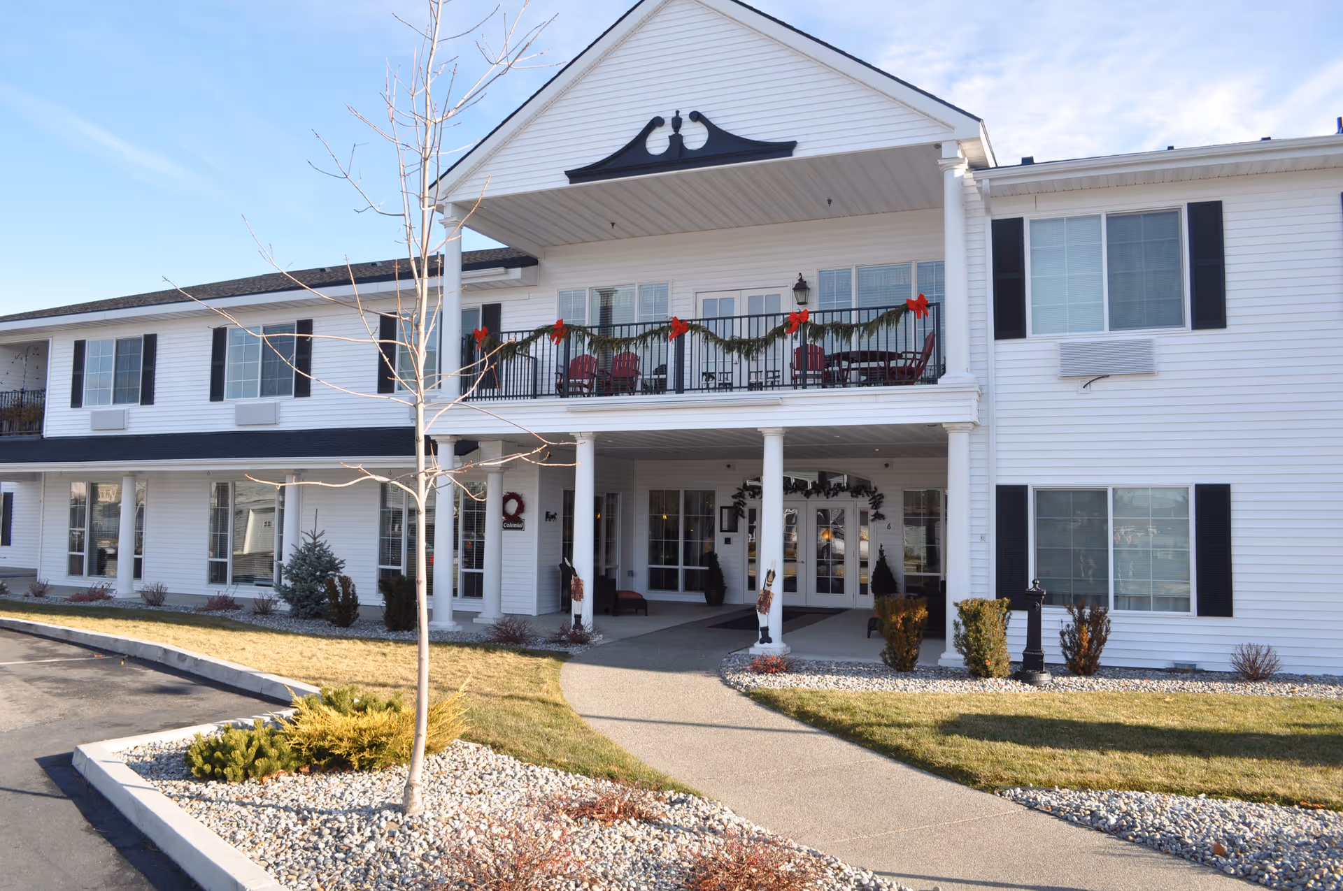 Front exterior view of a two-story white building with black shutters and a covered entrance supported by white columns. The entrance is decorated with garlands and red bows, and there is a balcony above with red chairs and similar decorations. The surrounding area has a paved walkway, small trees, and landscaped bushes.