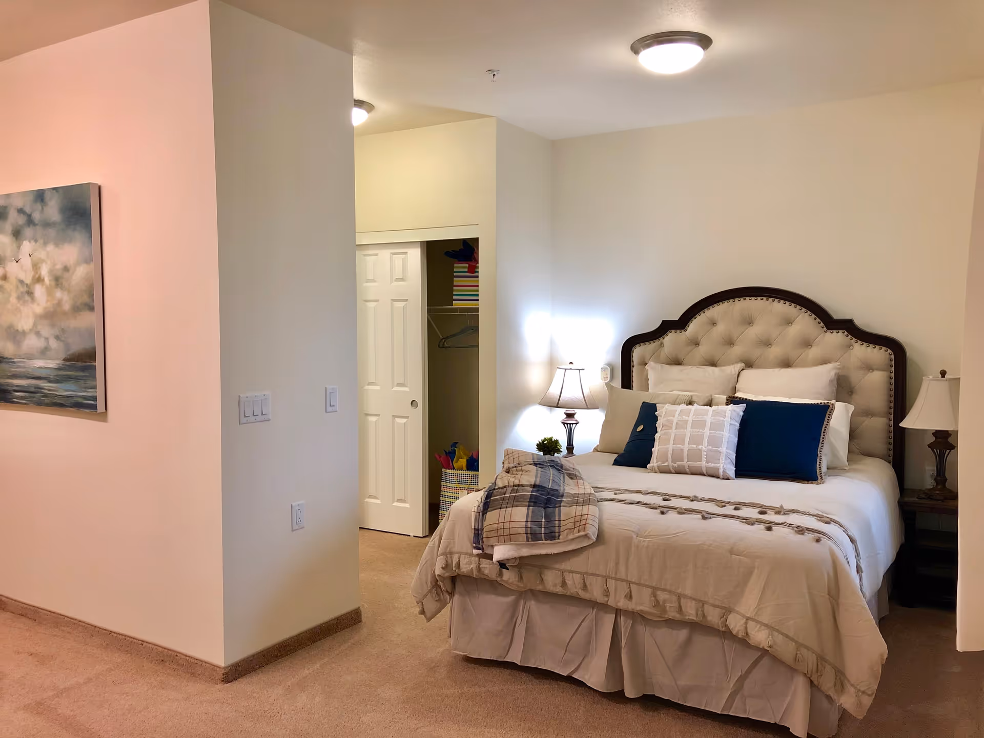 A cozy bedroom with a beige tufted headboard bed adorned with multiple pillows and a folded blanket. Two bedside tables each hold a lamp. There is a closet with sliding white doors partially open, revealing some colorful items inside. The walls are painted light beige, and a painting of a cloudy sky over water hangs on the left wall.