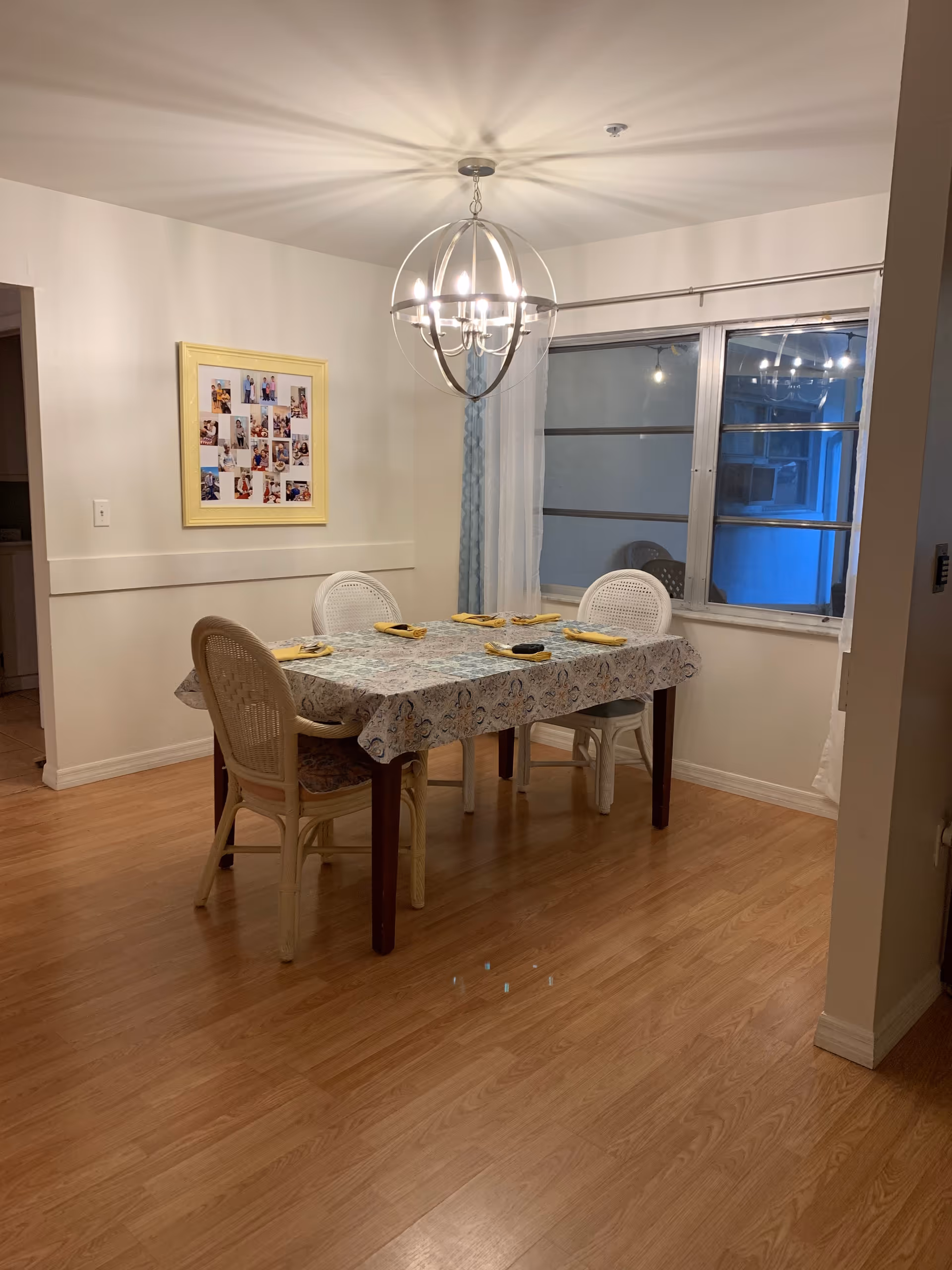A dining room with a rectangular table covered with a patterned tablecloth and set with yellow napkins. Four chairs surround the table. A modern chandelier hangs above the table, casting light patterns on the ceiling. There is a window with sheer curtains on one wall and a framed collage of photos on another wall. The floor is wooden.