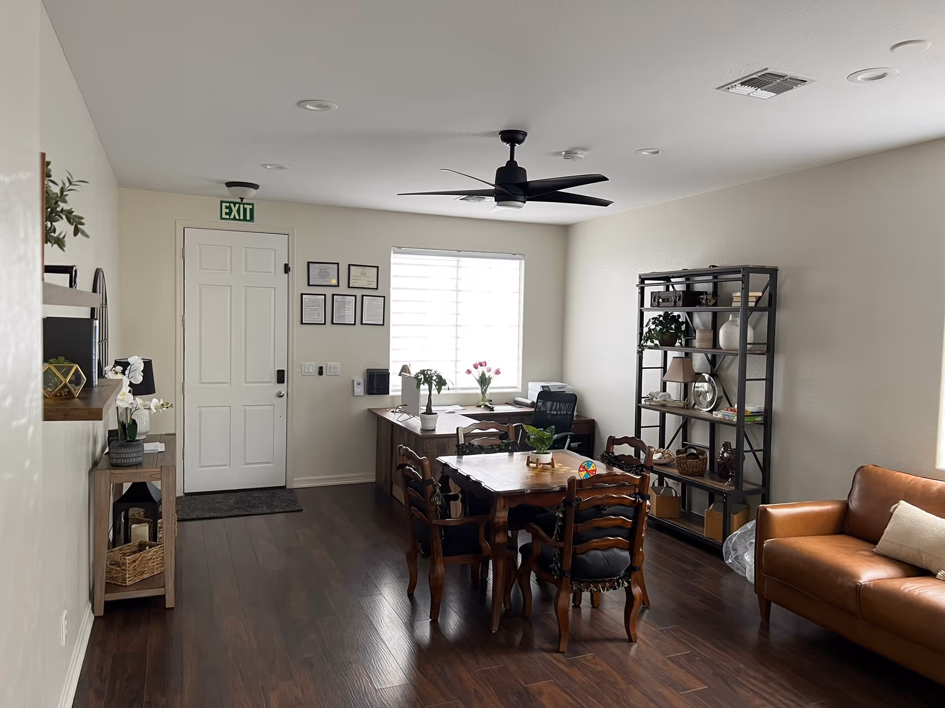 Bright interior common room with a wooden dining table and chairs, a desk by the window, a leather sofa, and shelving against the wall.