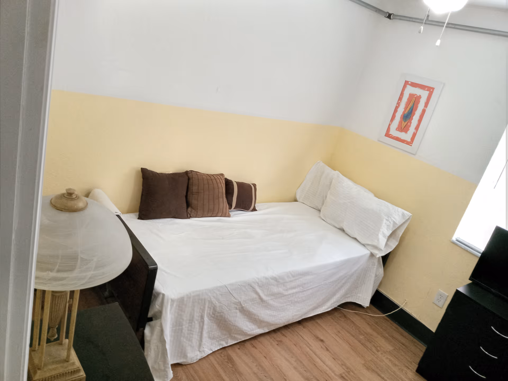 Small bedroom with a single bed against a two-tone yellow and white wall, several pillows, a bedside lamp and a TV on a cabinet.
