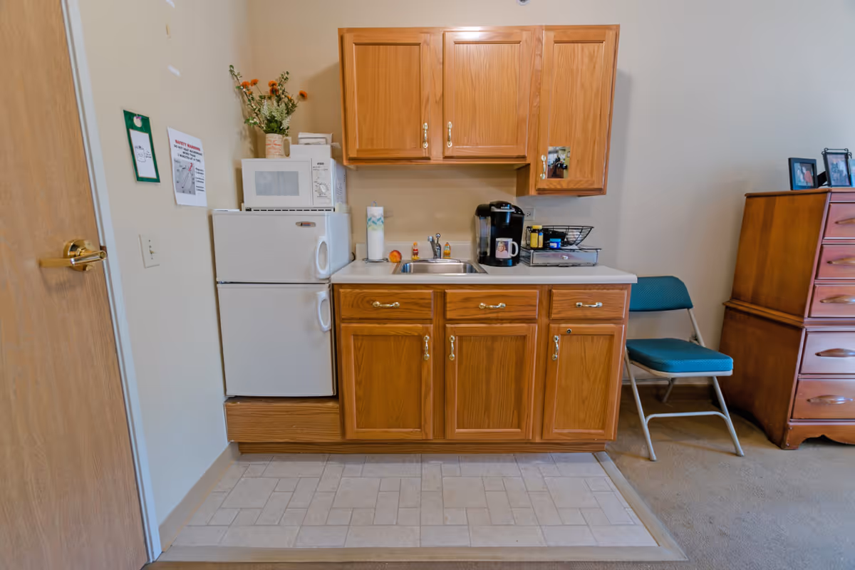 A small kitchenette area with wooden cabinets, a white mini refrigerator topped with a microwave, a sink, a coffee maker, paper towels, and some decorative items. To the right, there is a green folding chair and a wooden dresser with framed photos on top.