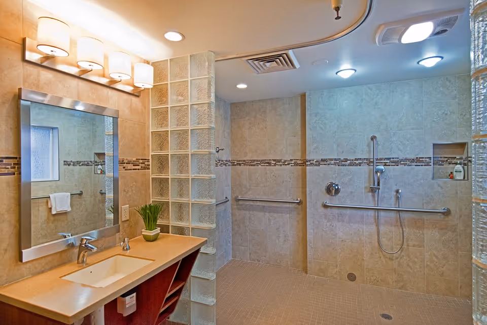 A spacious, accessible bathroom with beige tiled walls and floor. The bathroom features a large mirror above a countertop with a sink and a small potted plant. There is a walk-in shower area with grab bars and a handheld showerhead, separated by a glass block partition. The lighting includes wall-mounted fixtures above the mirror and recessed ceiling lights.