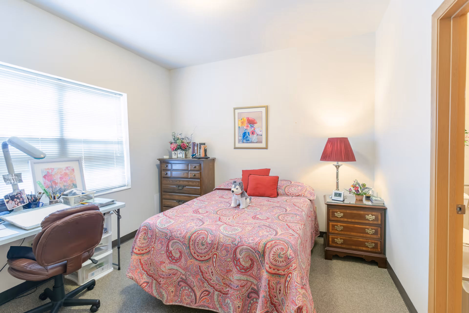 A cozy bedroom with a bed covered in a red and pink paisley patterned bedspread, two red pillows, and a small stuffed dog on top. To the right of the bed is a wooden nightstand with a red lampshade, a digital clock, and a small flower arrangement. On the left side of the room, there is a wooden dresser with books and flowers on top, and a white desk with a brown office chair in front of a window with blinds. The walls are light-colored and there is a framed floral picture hanging above the bed.
