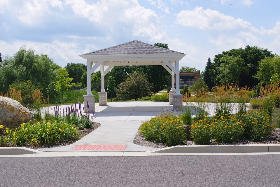 White pavilion/gazebo over a paved walkway with landscaped flower beds and trees under a partly cloudy sky.
