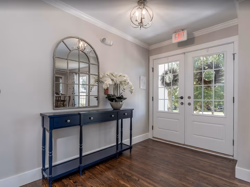 Bright entry foyer with double glass doors, a navy console table topped with an orchid and an arched mirror, and hardwood floors.