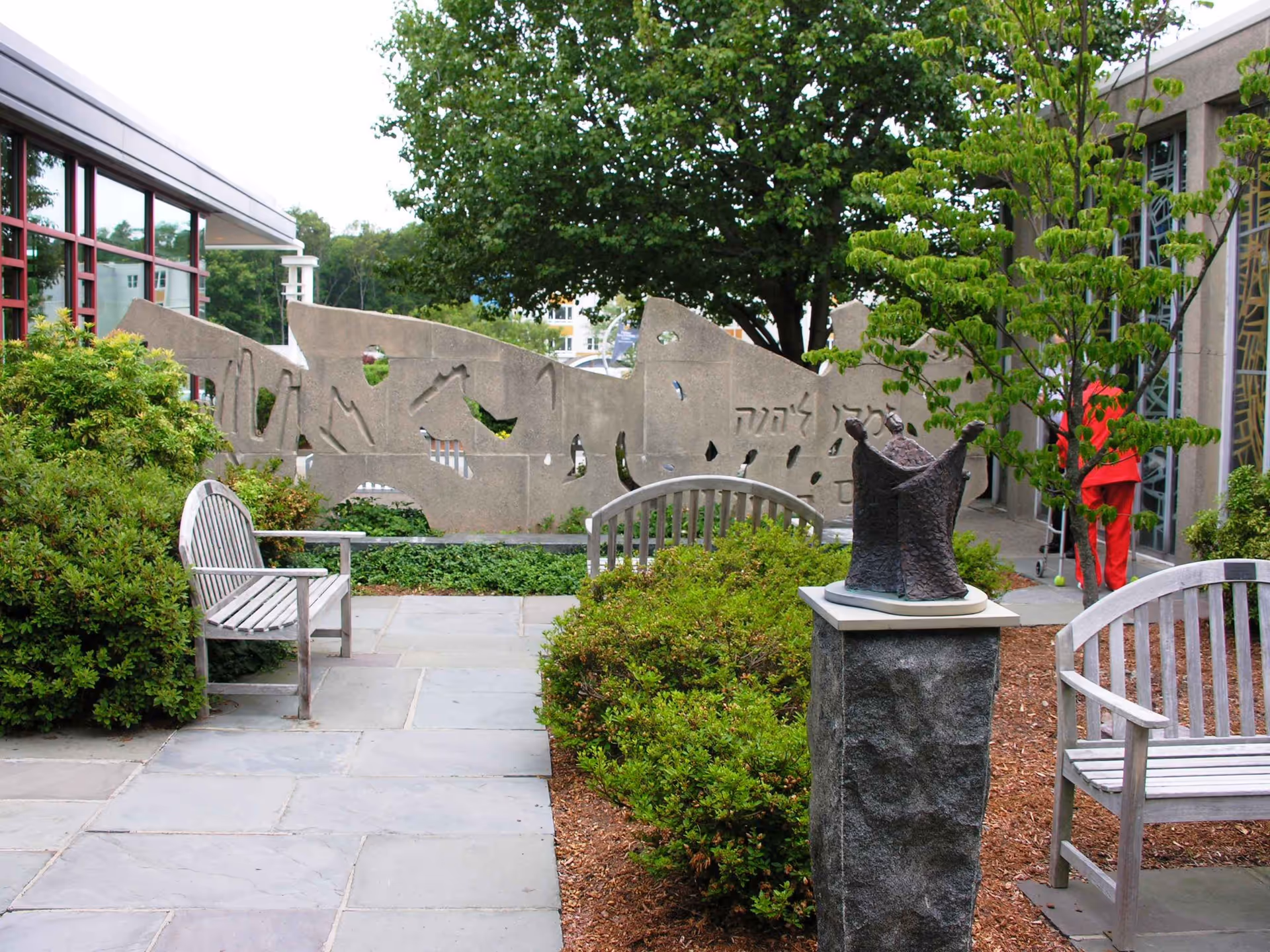 Outdoor garden area at Hebrew Rehabilitation Center - Boston with stone benches, green bushes, a tree, a stone sculpture on a pedestal, and a decorative stone wall with Hebrew inscriptions. A person in red clothing is visible near the building entrance.