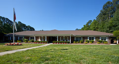 Single-story brick building with a brown roof, surrounded by green grass and trees under a clear blue sky. There is a flagpole with an American flag and another flag in front of the building, along with flower beds lining the walkway leading to the entrance.