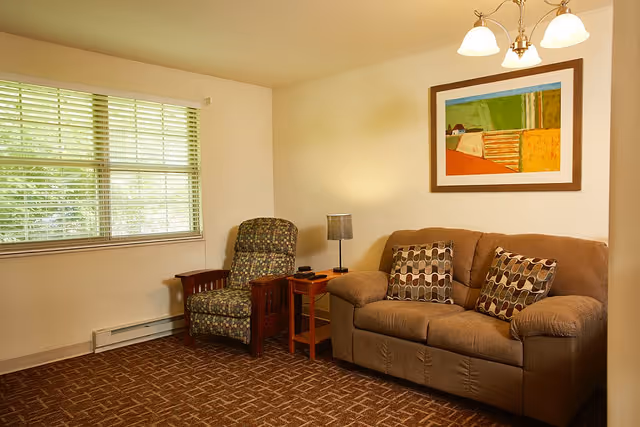 A cozy living room with a brown sofa adorned with patterned cushions, a cushioned armchair with a wooden frame, a small wooden side table with a lamp, a large window with blinds, and a colorful framed painting on the wall.
