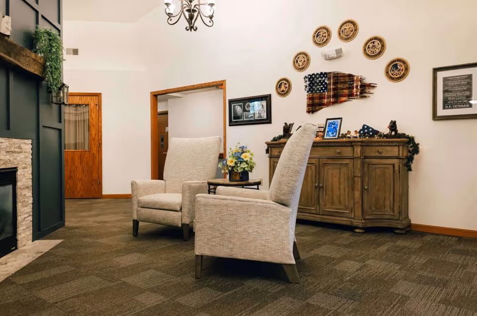 Cozy interior lounge with two upholstered armchairs around a small table, a wooden sideboard with patriotic wall decor, and a fireplace.