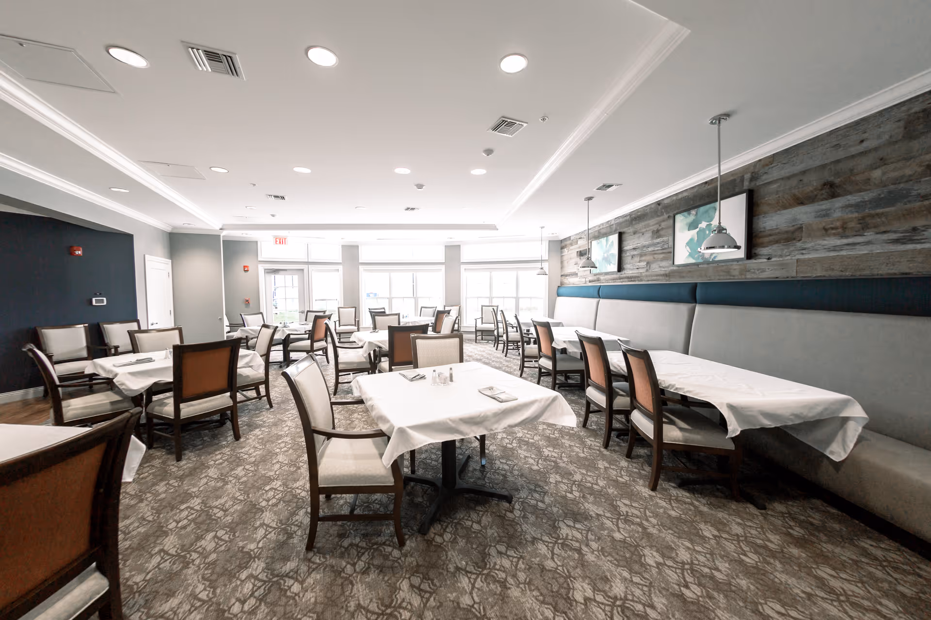 Bright and spacious dining room with multiple tables covered in white tablecloths, surrounded by wooden chairs with cushioned seats. The room features large windows letting in natural light, a patterned carpet, and a wooden accent wall with framed artwork and hanging pendant lights.