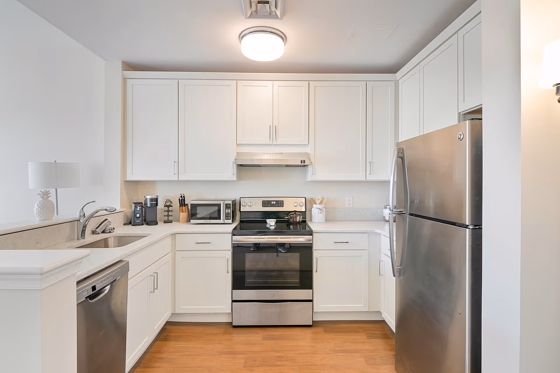 A modern kitchen with white cabinets, stainless steel appliances including a refrigerator, oven, and dishwasher. The kitchen has a microwave, coffee maker, knife block, and a small lamp on the counter. The floor is wooden, and the walls are painted white.