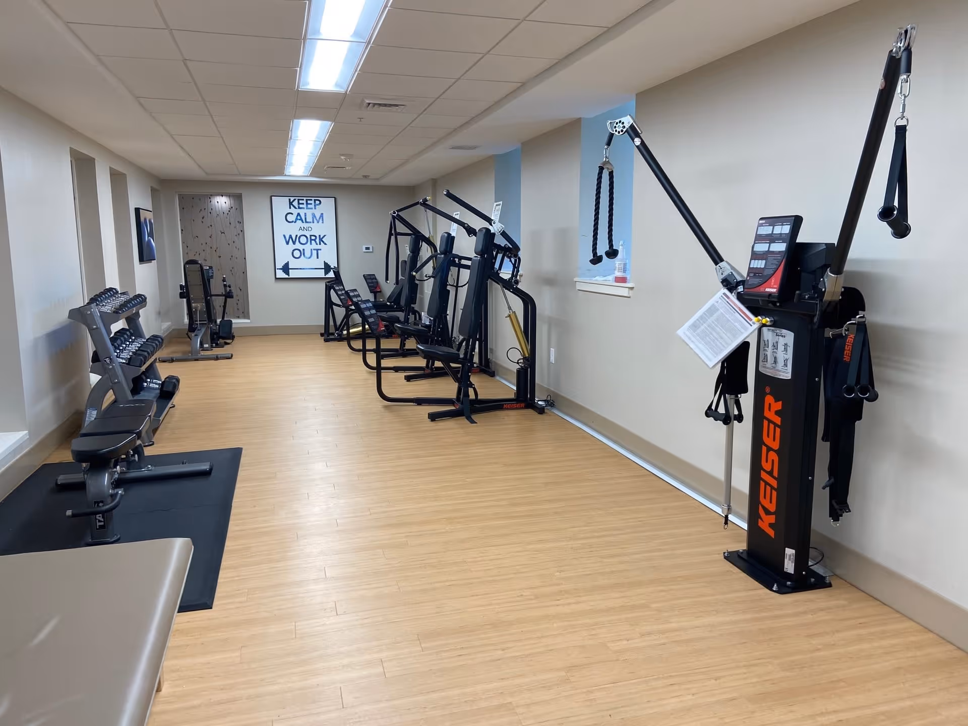 Interior view of a fitness room with various exercise machines and equipment including weight benches, dumbbells, and resistance machines. A motivational sign on the wall reads 'KEEP CALM AND WORK OUT'. The room has light-colored walls, wood flooring, and overhead fluorescent lighting.