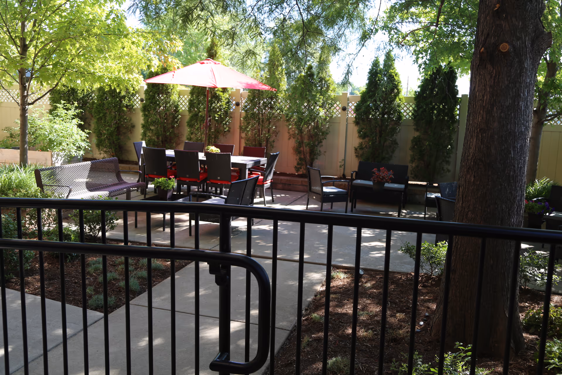 Outdoor patio area with a table and chairs under a red umbrella, surrounded by greenery and trees. There are additional seating areas with benches and chairs, and a black metal fence in the foreground.