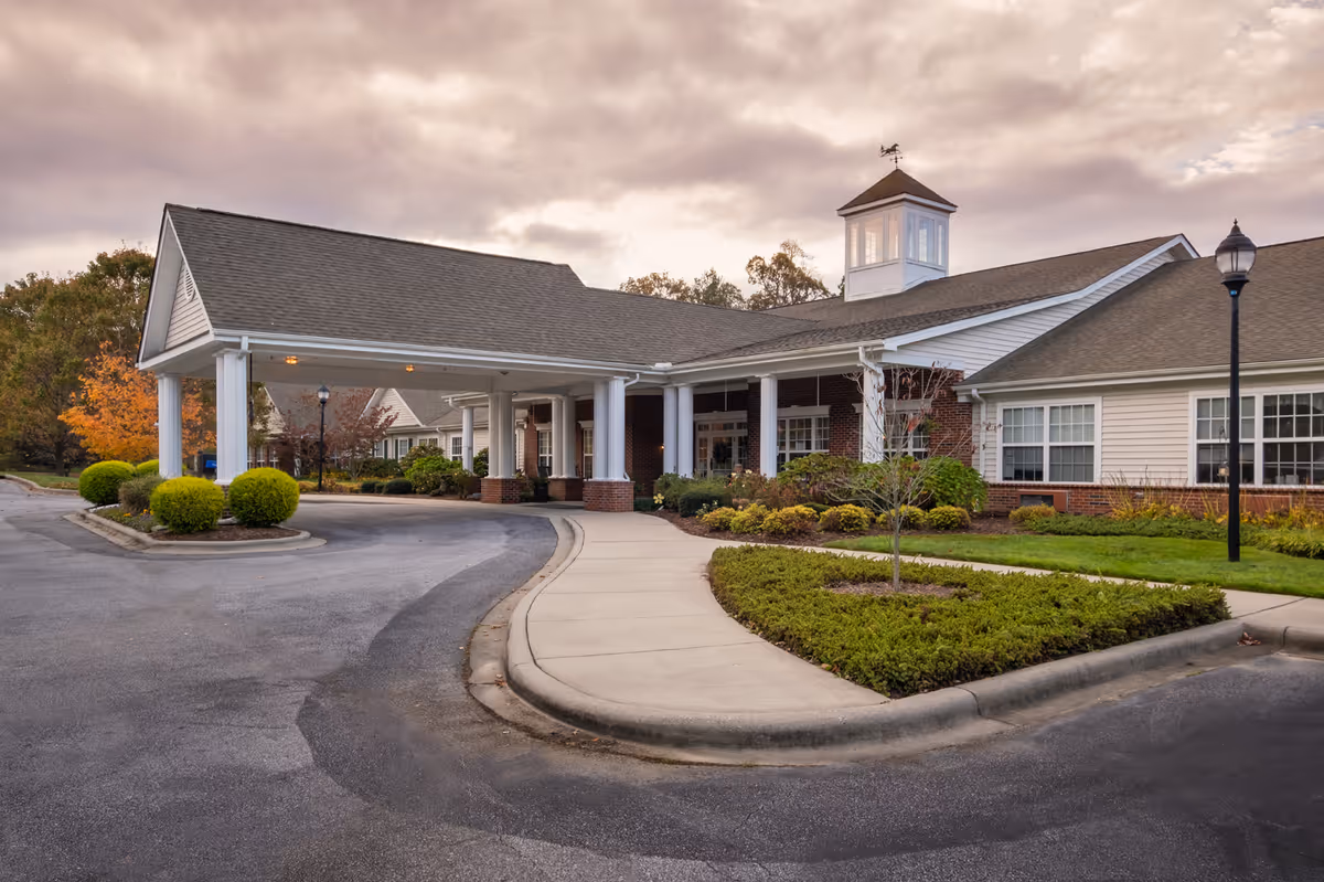Exterior view of a senior living facility building with a covered entrance, white columns, brick and siding walls, landscaped bushes, and a curved driveway under a cloudy sky.