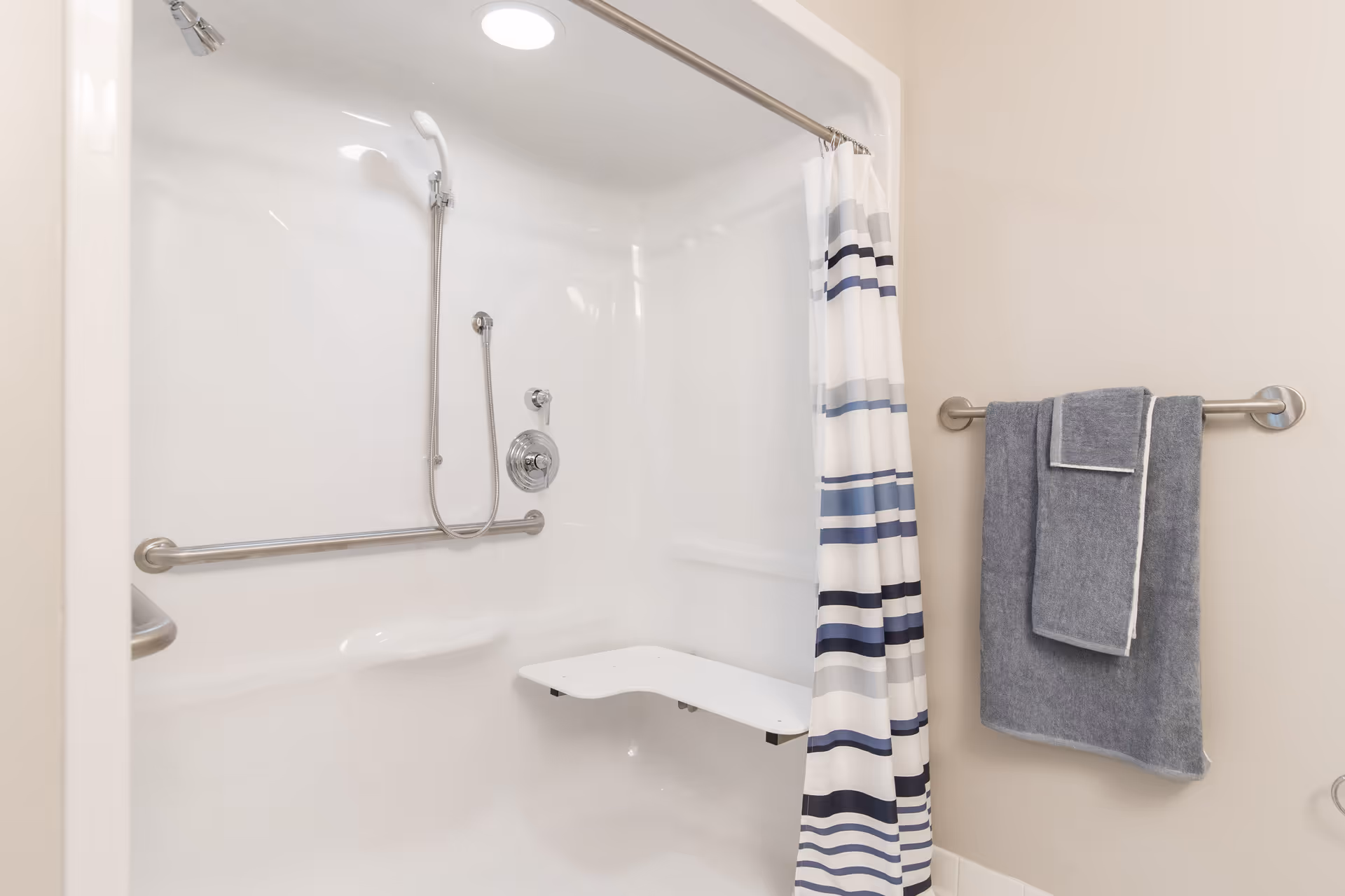 A clean, white shower area with a built-in foldable seat, a handheld showerhead, a horizontal grab bar, and a shower curtain with blue, gray, and black stripes. A towel rack on the adjacent wall holds two gray towels.