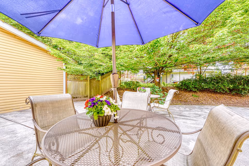 Outdoor patio area with a round metal table and four chairs. A large blue umbrella is open above the table, and a small flower pot with colorful flowers is placed on the table. Surrounding the patio are green trees and bushes, with a wooden fence and a yellow building visible in the background.