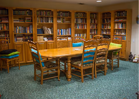 A cozy library room with wooden bookshelves filled with books lining the walls. In the center, there is a wooden table surrounded by six wooden chairs, each with a colorful cushion. The floor is carpeted with a patterned green carpet, and the room is warmly lit.