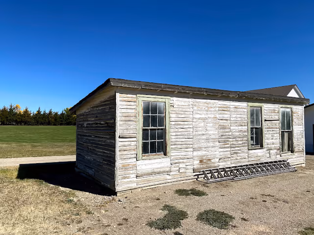 An old, weathered wooden building with peeling white paint and three windows. A long metal ladder is lying on the ground against the side of the building. The building is situated on a dirt and grass area with a clear blue sky overhead and a line of trees in the background.