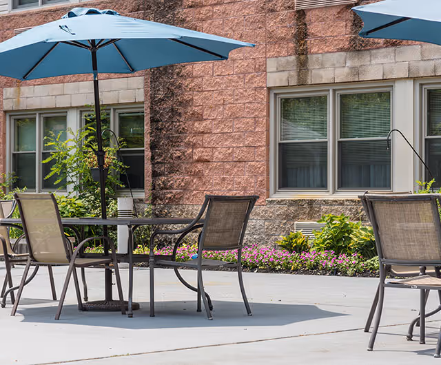 Patio with tables, chairs, and blue umbrellas outside a brick building with windows and flower beds.