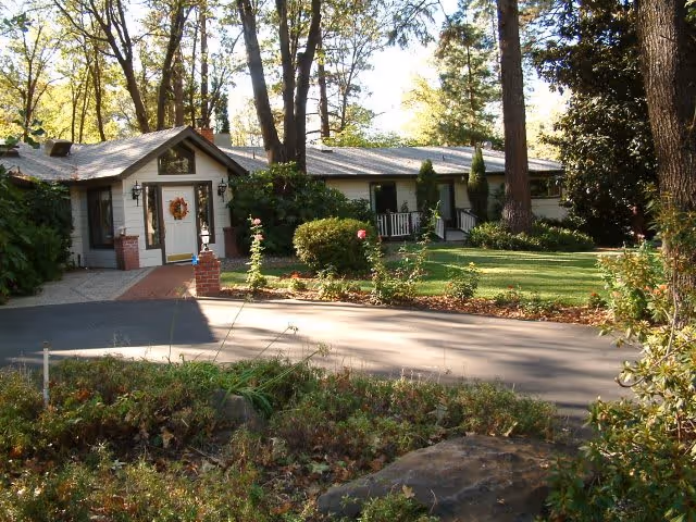 Single-story residential building surrounded by trees and greenery with a paved driveway and a well-maintained lawn in front. The entrance has a small porch with a wreath on the door.
