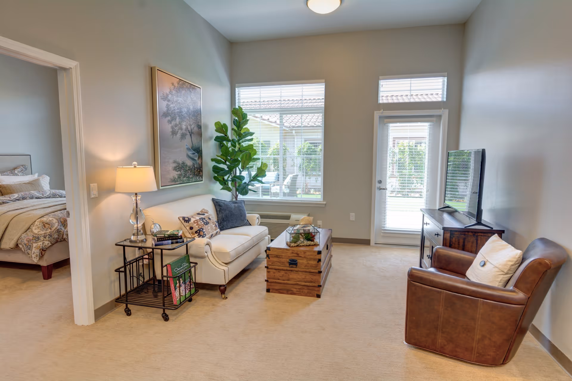 A cozy living room in Rose Arbor Village featuring a white sofa with decorative pillows, a wooden chest used as a coffee table, a brown leather armchair with a pillow, a TV on a wooden stand, a side table with a lamp and books, a large plant near the window, and a view into a bedroom with a made bed.
