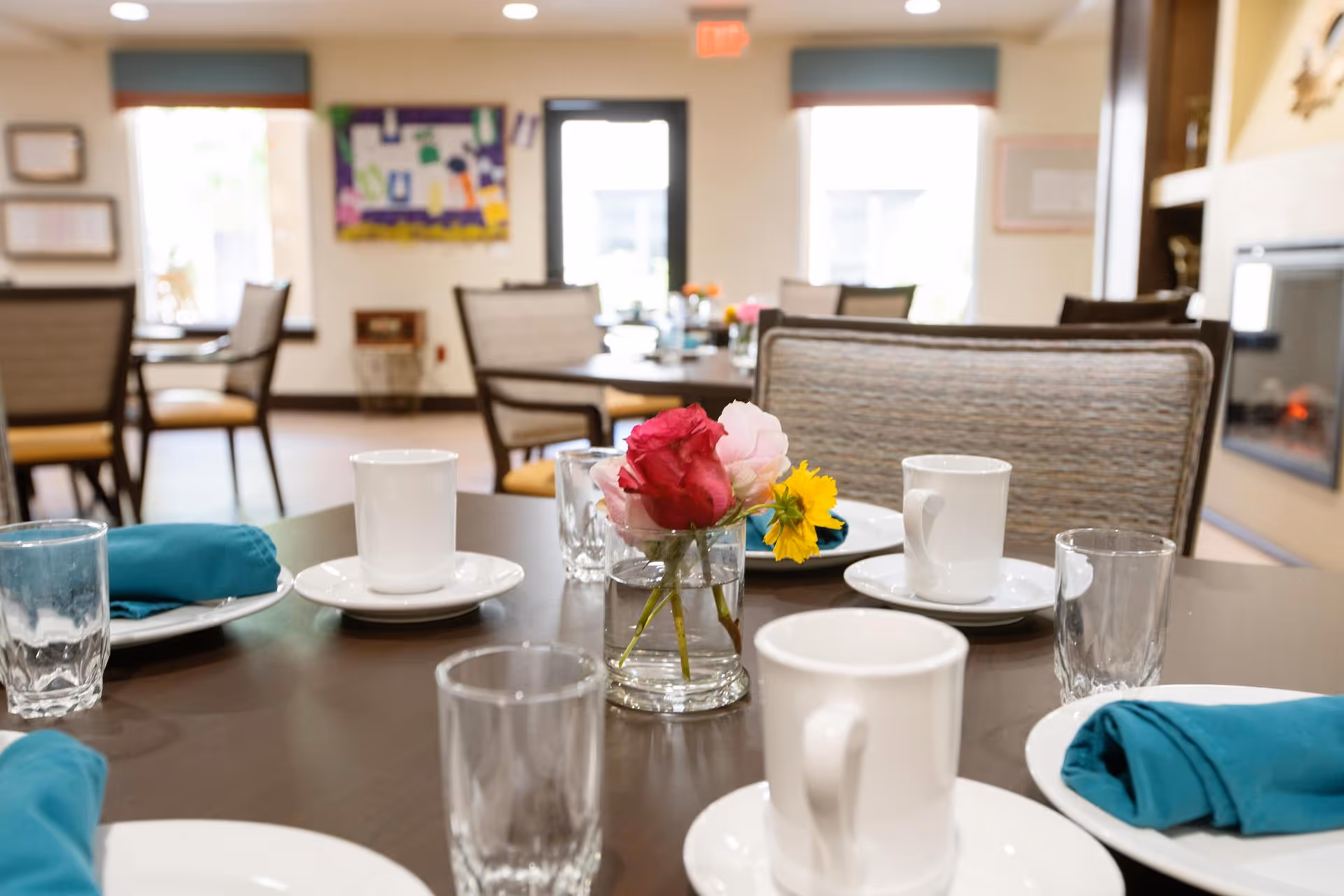 Dining table set with white cups, glasses, teal napkins and a small vase of flowers in a bright communal dining room.