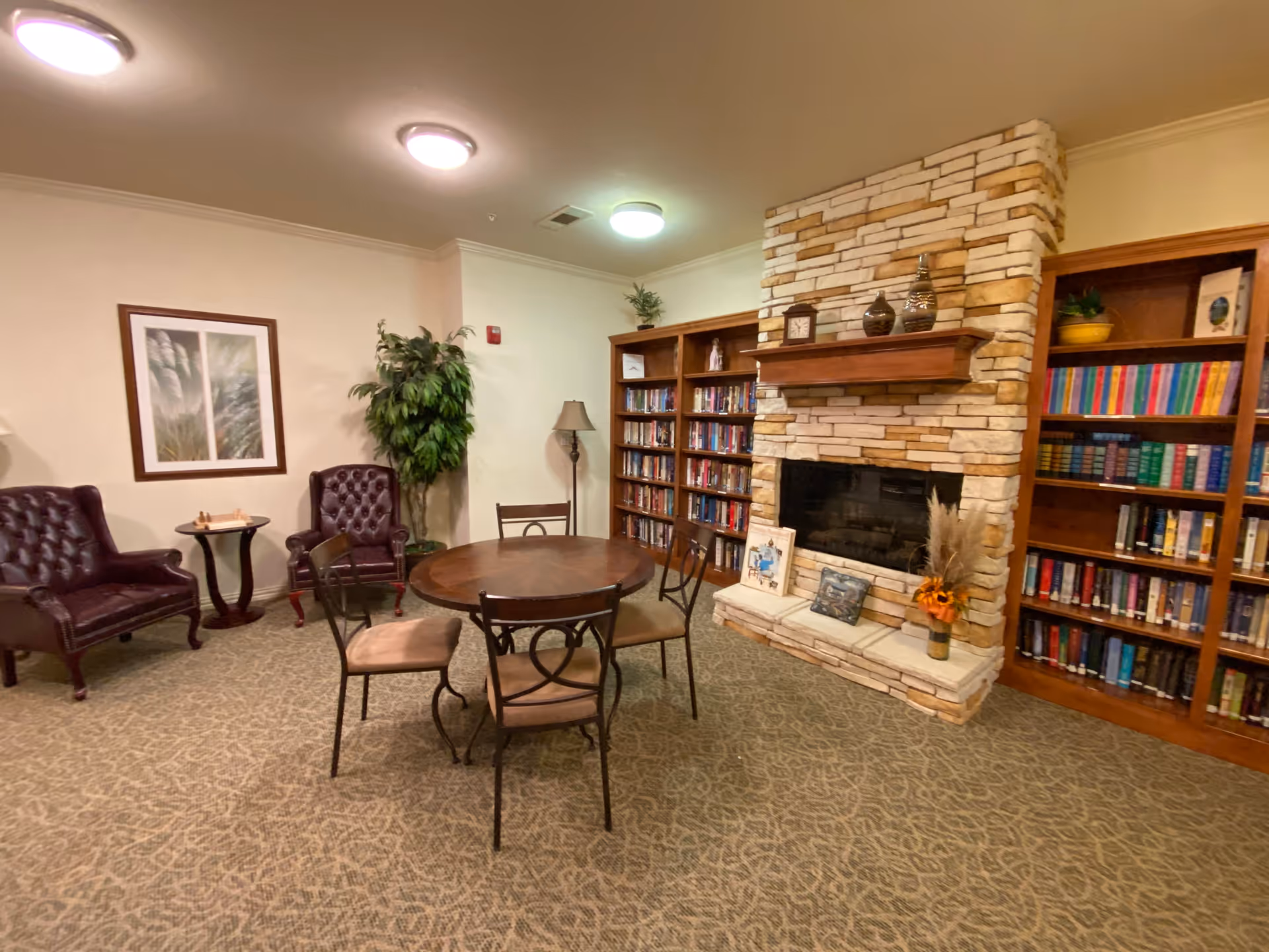 A cozy common area with a round wooden table surrounded by four chairs in the center. To the right, there is a stone fireplace with decorative items on the mantel and a small vase with flowers on the hearth. Behind the fireplace are two wooden bookshelves filled with books. On the left side, there are two dark leather armchairs with a small round table between them, a framed picture on the wall, a tall green plant, and a floor lamp. The room has beige walls and carpeted flooring with a patterned design.