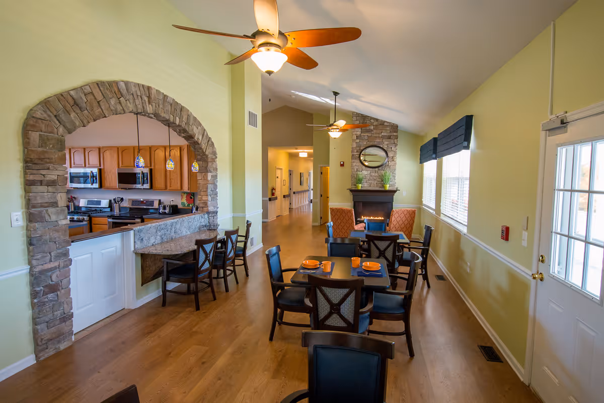 Interior view of a senior living facility dining area with wooden floors, several tables and chairs set with orange plates and mugs, a stone archway leading to a kitchen with wooden cabinets and stainless steel appliances, ceiling fans with lights, and a stone fireplace with a round mirror above it.