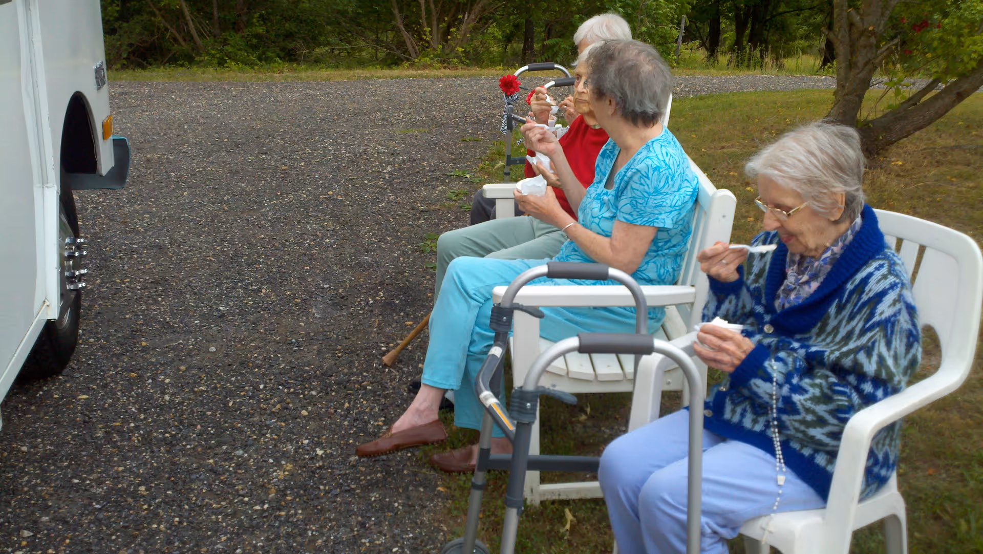 Four elderly women sitting outdoors on white chairs, eating from small bowls with spoons. Two walkers are positioned near the women. They are seated on a grassy area next to a gravel path with trees in the background.
