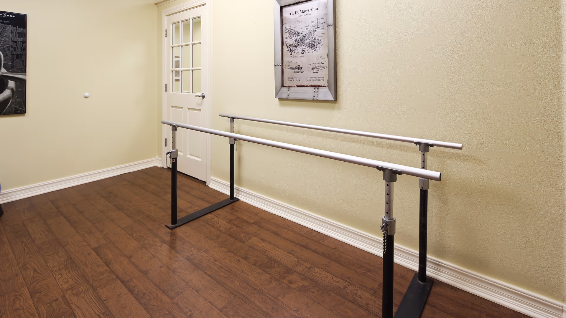 A room with wooden flooring and light yellow walls featuring a set of parallel metal handrails used for walking or physical therapy. A white door with glass panels is visible in the background, along with framed artwork on the walls.
