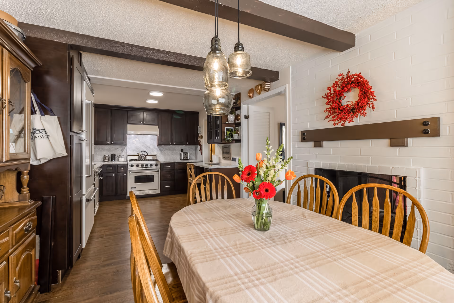 Dining area with a vase of red and orange flowers on a plaid-covered table, wooden chairs, a fireplace on the right, and a dark-wood kitchen visible in the background.