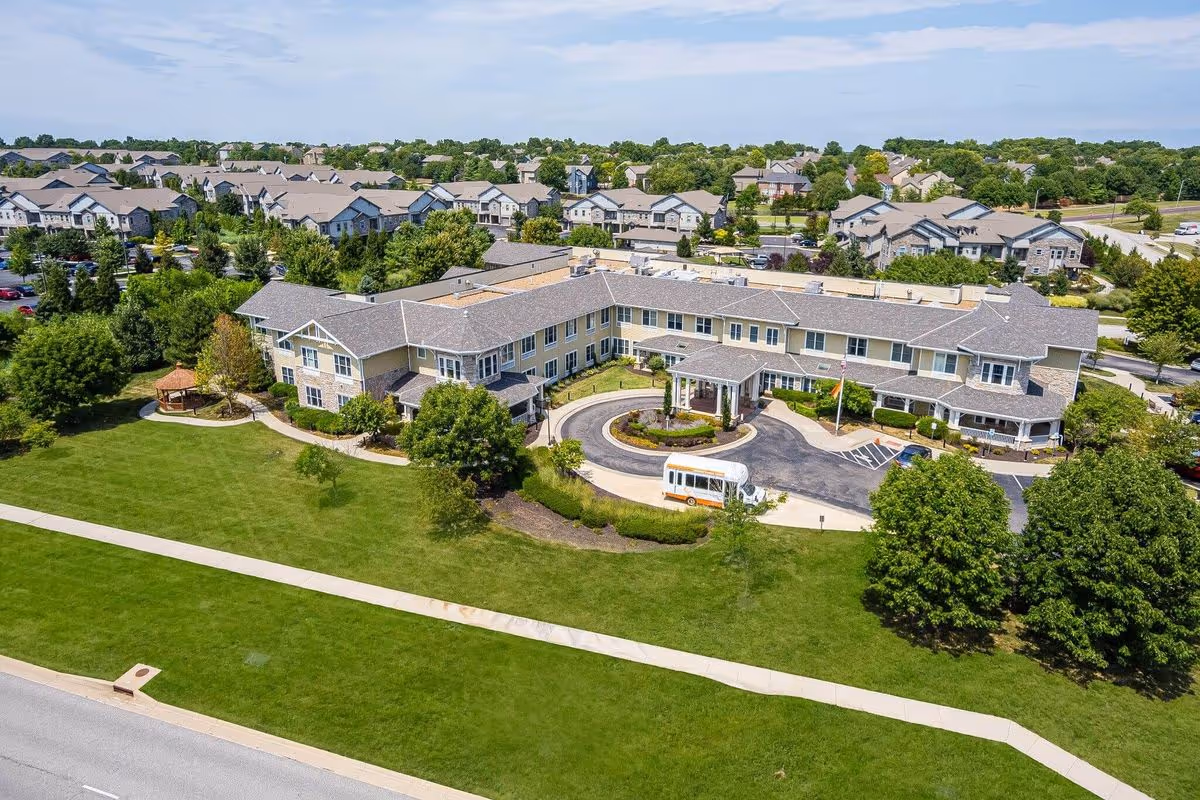 Aerial view of Sunrise of Overland Park senior living facility showing a large two-story building with a circular driveway and a small shuttle bus parked in front. The building is surrounded by well-maintained green lawns, trees, and a gazebo on the left side. Residential houses and streets are visible in the background under a partly cloudy sky.