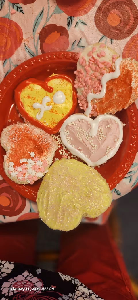A red plate holding several heart-shaped frosted cookies in pink, yellow, and red on a floral tablecloth.