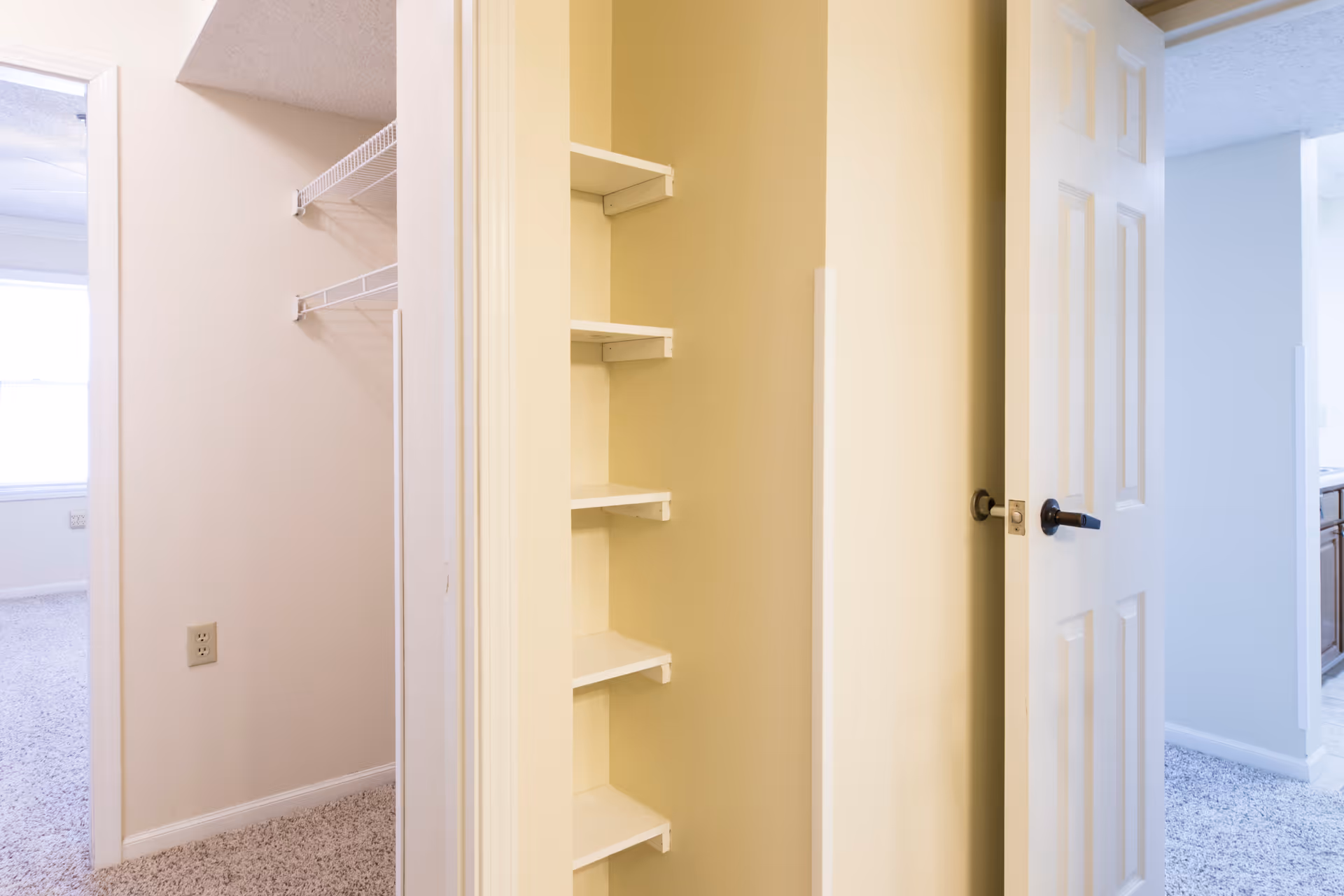 Interior view of a hallway in a residential facility showing an open door with built-in shelves inside a small closet. To the left, there is a walk-in closet with wire shelving and a carpeted floor. The hallway leads to other rooms with light-colored walls and carpeted floors.