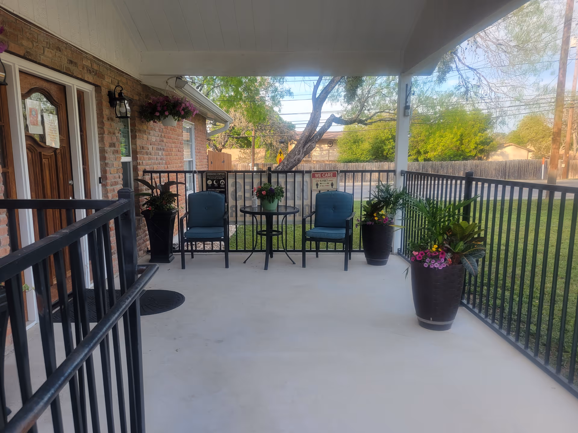 Covered outdoor patio area with two blue cushioned chairs and a small round table between them. The patio is enclosed with black metal railings and decorated with several large potted plants with flowers. A wooden door and brick wall are visible on the left side, and a grassy yard with trees and a wooden fence is in the background.