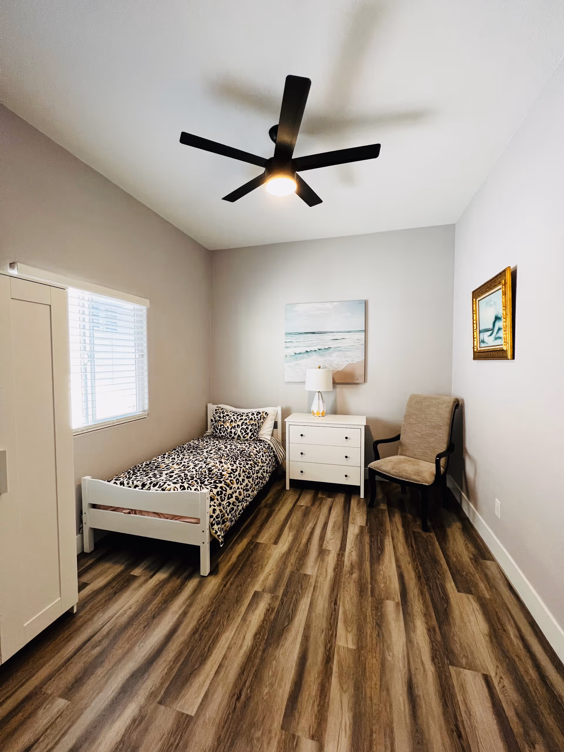 A small, neatly arranged bedroom with a single bed featuring leopard print bedding, a white nightstand with a lamp, a beige upholstered chair, a white wardrobe, and two framed pictures on the walls. The room has wood-look flooring and a modern black ceiling fan with a light.