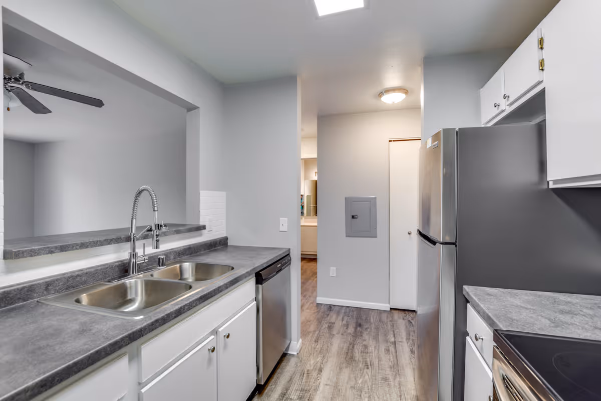 Modern kitchen with gray countertops, a double stainless steel sink with a high-arc faucet, white cabinets, a stainless steel dishwasher, refrigerator, and stove, and wood-look flooring. The kitchen opens to an adjacent room with a ceiling fan visible.