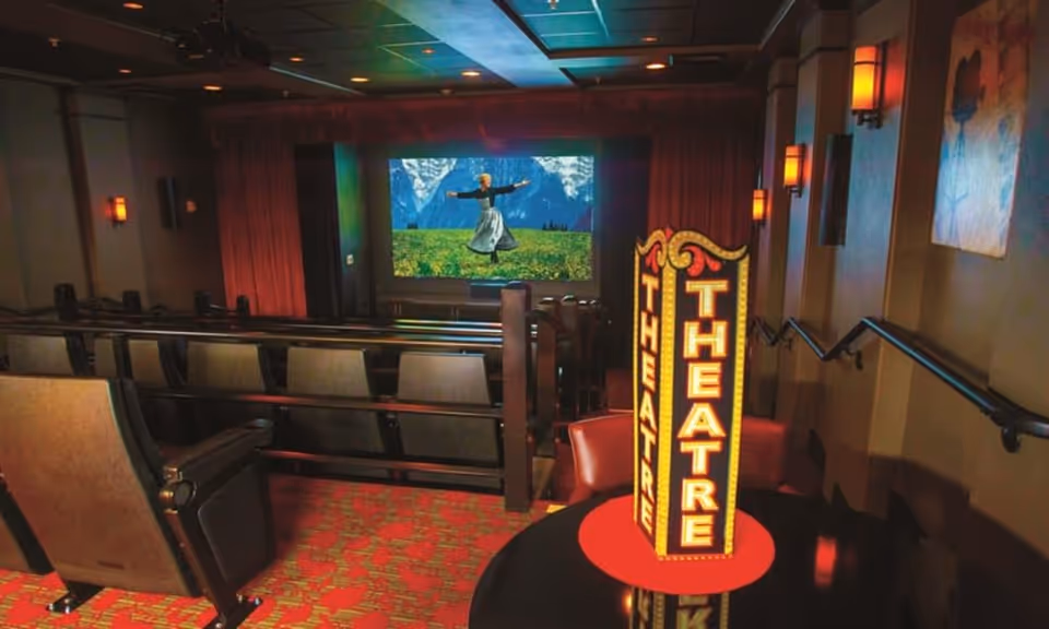 Interior view of a small theater room with rows of cushioned seats facing a large screen showing a scene from a movie with a woman in a dress standing in a field with mountains in the background. A decorative illuminated sign reading 'THEATRE' is placed on a round table near the front row. The room has warm lighting and wall sconces.